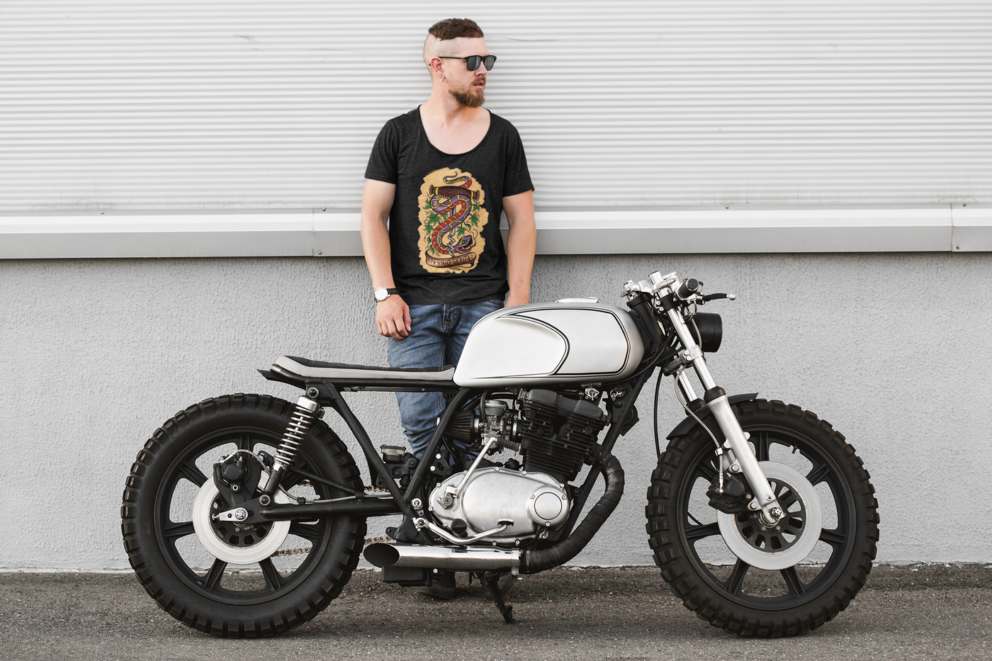 Hipster with punk hair cut, sunglasses and jeans standing next to a custom retro cafe racer motorcycle wearing jeans and a Vino Tees T shirt.